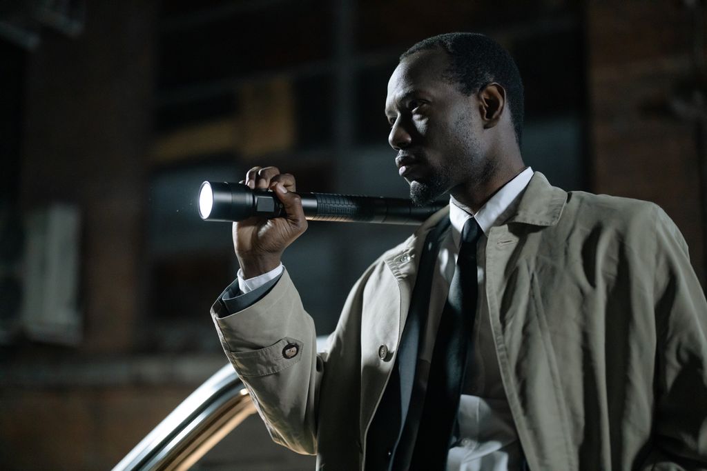 African-American detective in a beige trench coat holding a flashlight outdoors at night.